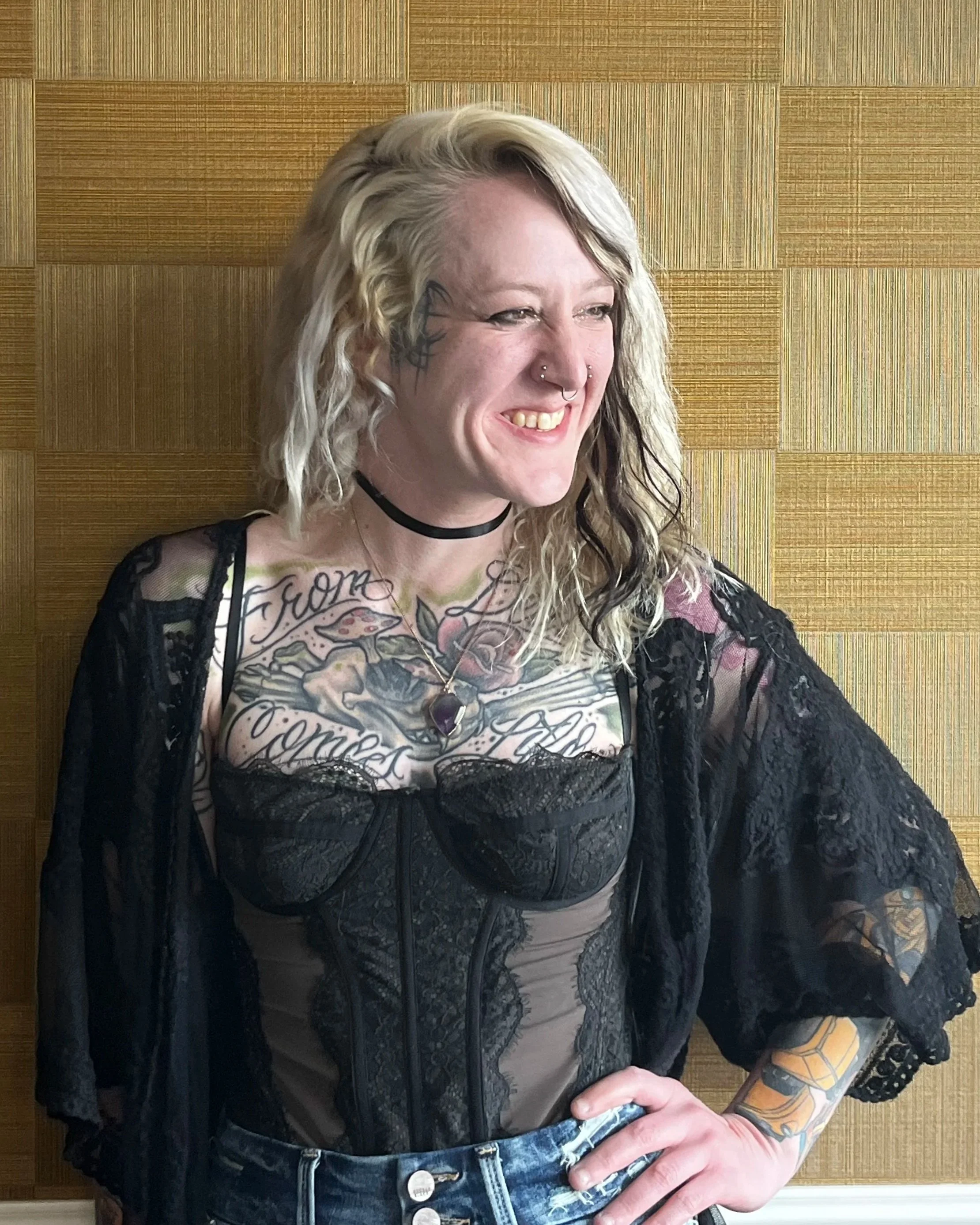 Young woman with blonde and black curly hair, tattoos on chest and arms, smiling, wearing a black lace corset top, mesh cover-up, denim jeans, in front of wooden wall.