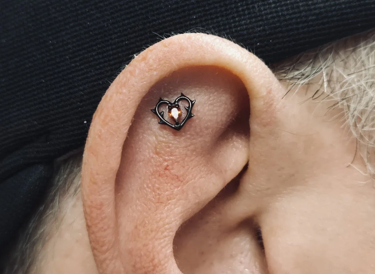 A close-up of a person’s ear with a black heart-shaped jewelry piece featuring a small gem.