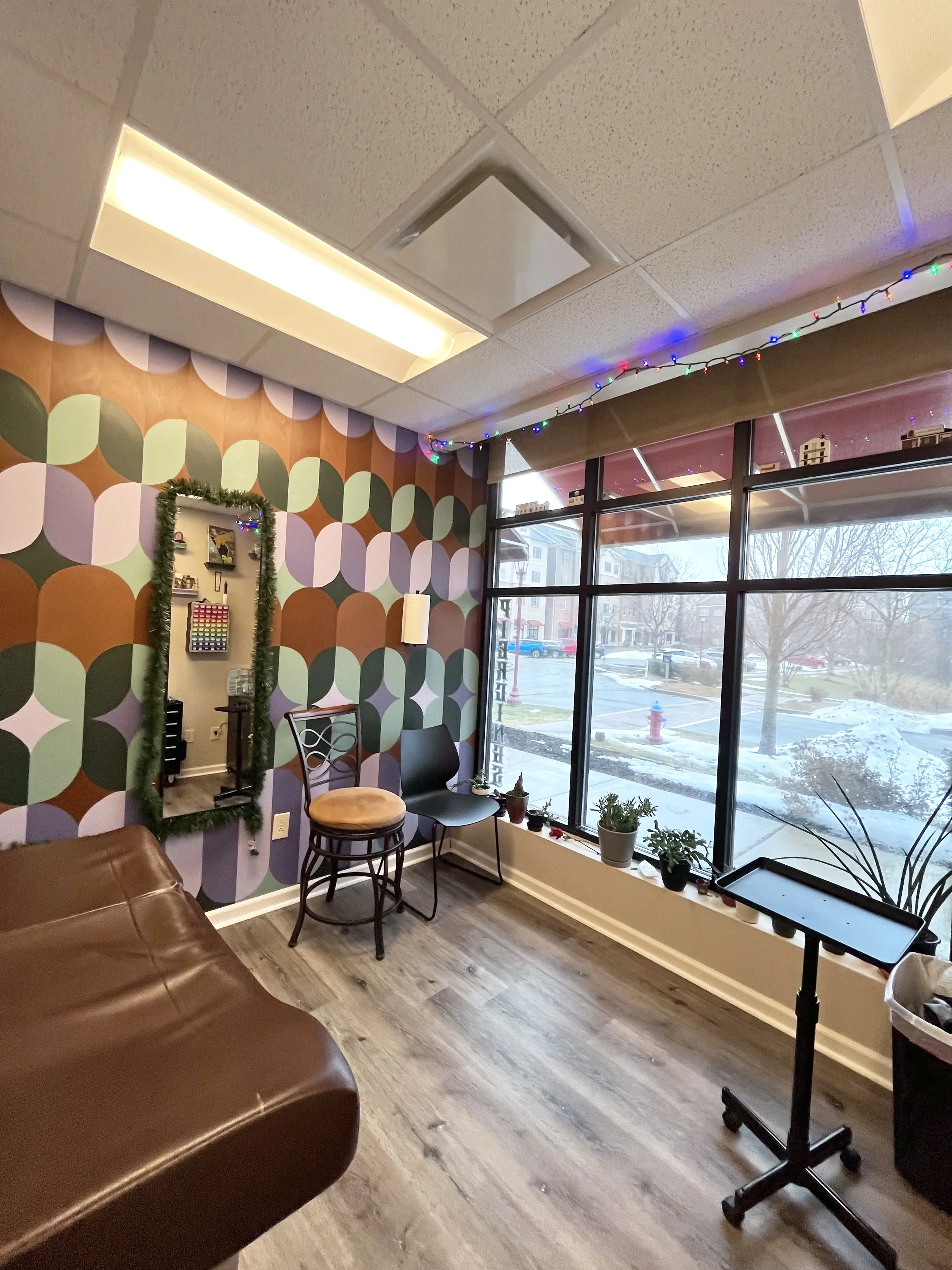 A cozy room with colorful circular-patterned wallpaper, a window with potted plants on the windowsill, chairs, a mirror decorated with a Christmas garland, and string lights hanging from the ceiling.