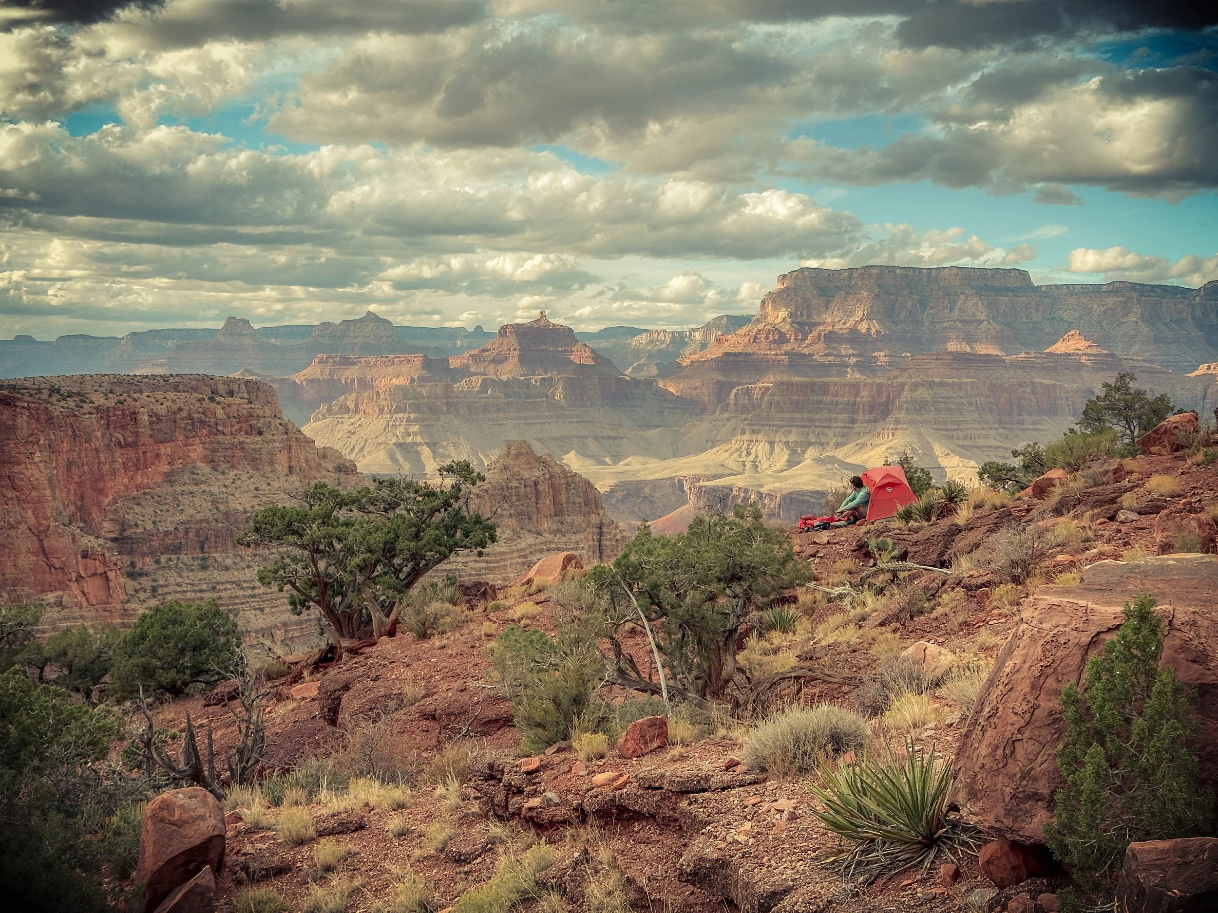 A person sitting outside a small red tent on the edge of the Grand Canyon, with layered rock formations and a cloudy sky in the background.