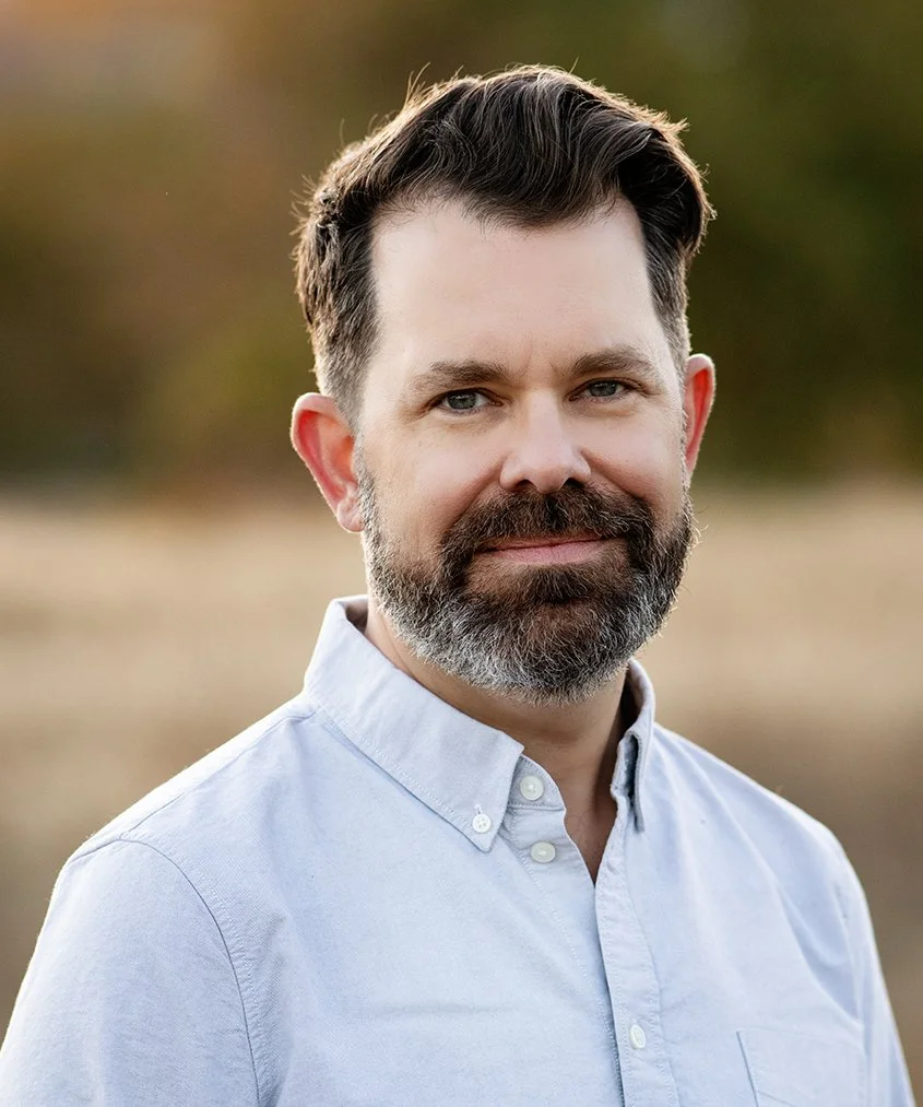 A portrait of Jordan Williams, Principal at Loop Trails Strategies. He has a beard and short dark hair, is wearing a white button-up shirt, and is standing outdoors against a blurred natural background.