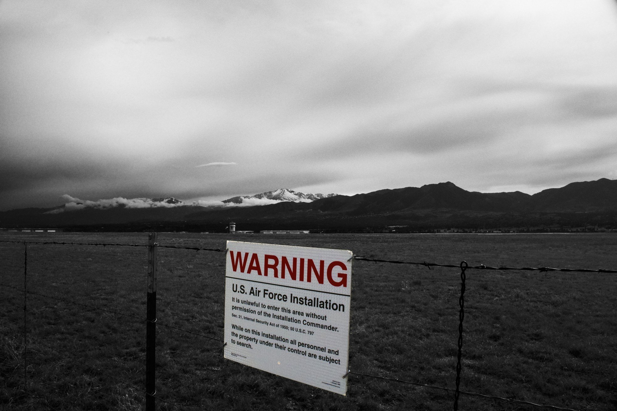 A black and white landscape with mountains in the background and a cloudy sky. There is a warning sign attached to a barbed wire fence in the foreground, indicating a U.S. Air Force installation, with restrictions on trespassing.