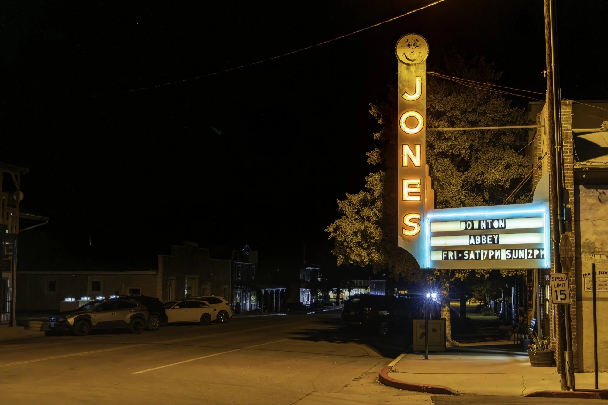 Night view of a vintage neon sign for Jones theater with a smiling face logo, advertising shows in Downtown on a marquee with scheduling details, illuminated by streetlights, with parked cars and buildings in the background.