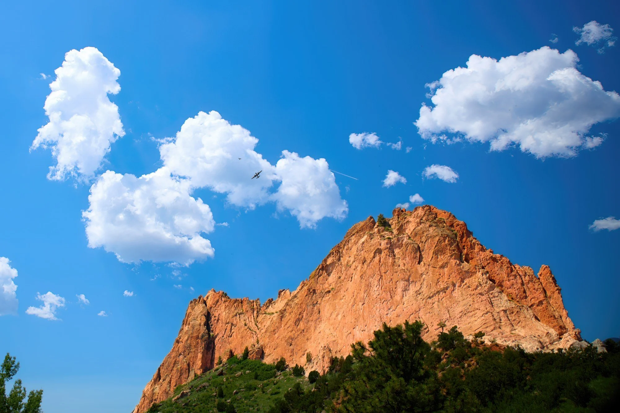A rugged red rock mountain with pine trees at its base, set against a bright blue sky with scattered white clouds and a small airplane flying in the distance.