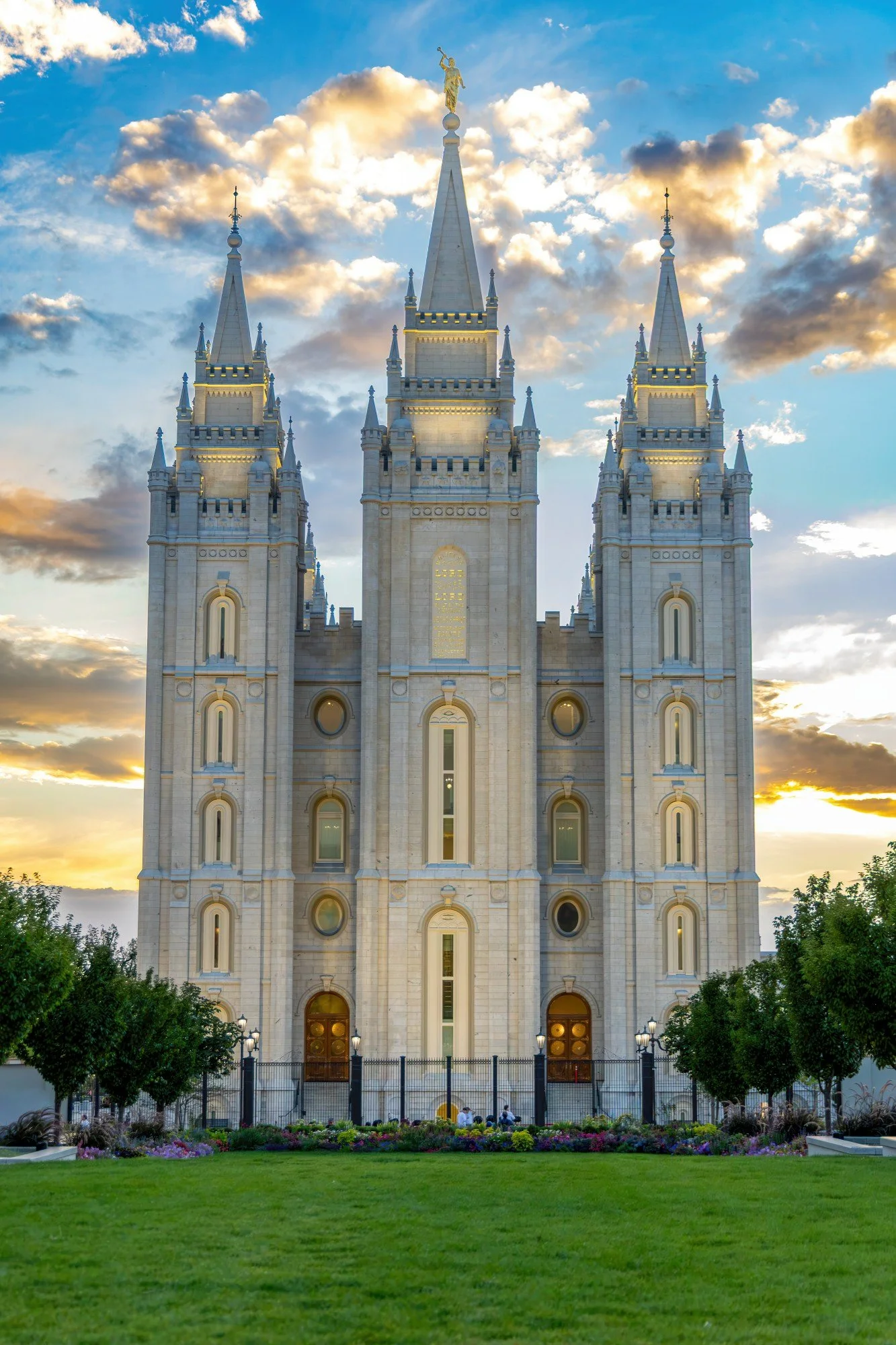 The image shows a tall, ornate white church or cathedral with a central spire topped with a golden angel statue, set against a partly cloudy sky during the sunset. The building has multiple pointed spires and decorative architectural details, with a fenced entrance and a well-maintained grassy lawn and flower beds in the foreground.