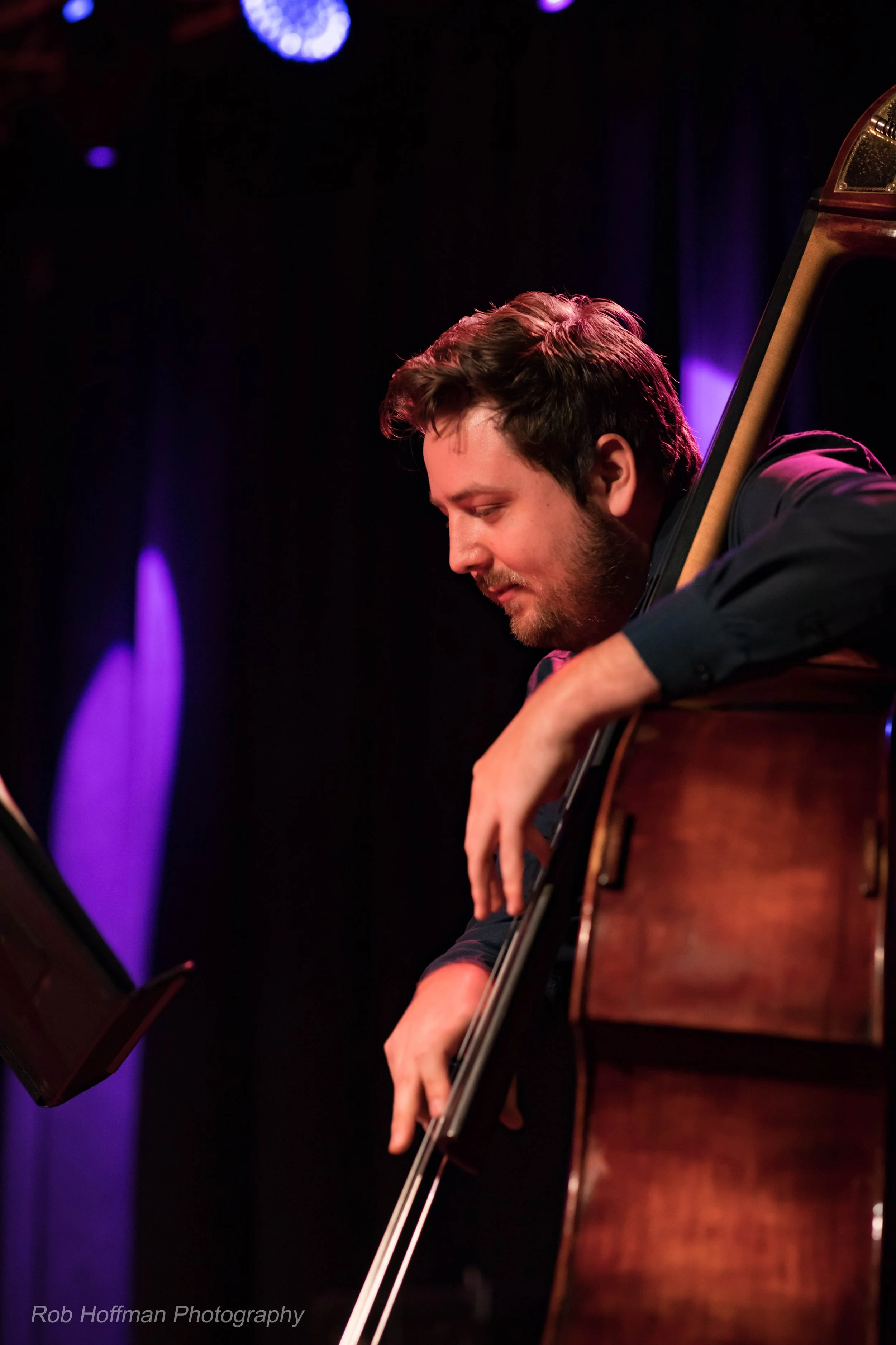 A young man with dark hair and a beard playing a guitar on stage, looking down, with purple stage lights in the background.