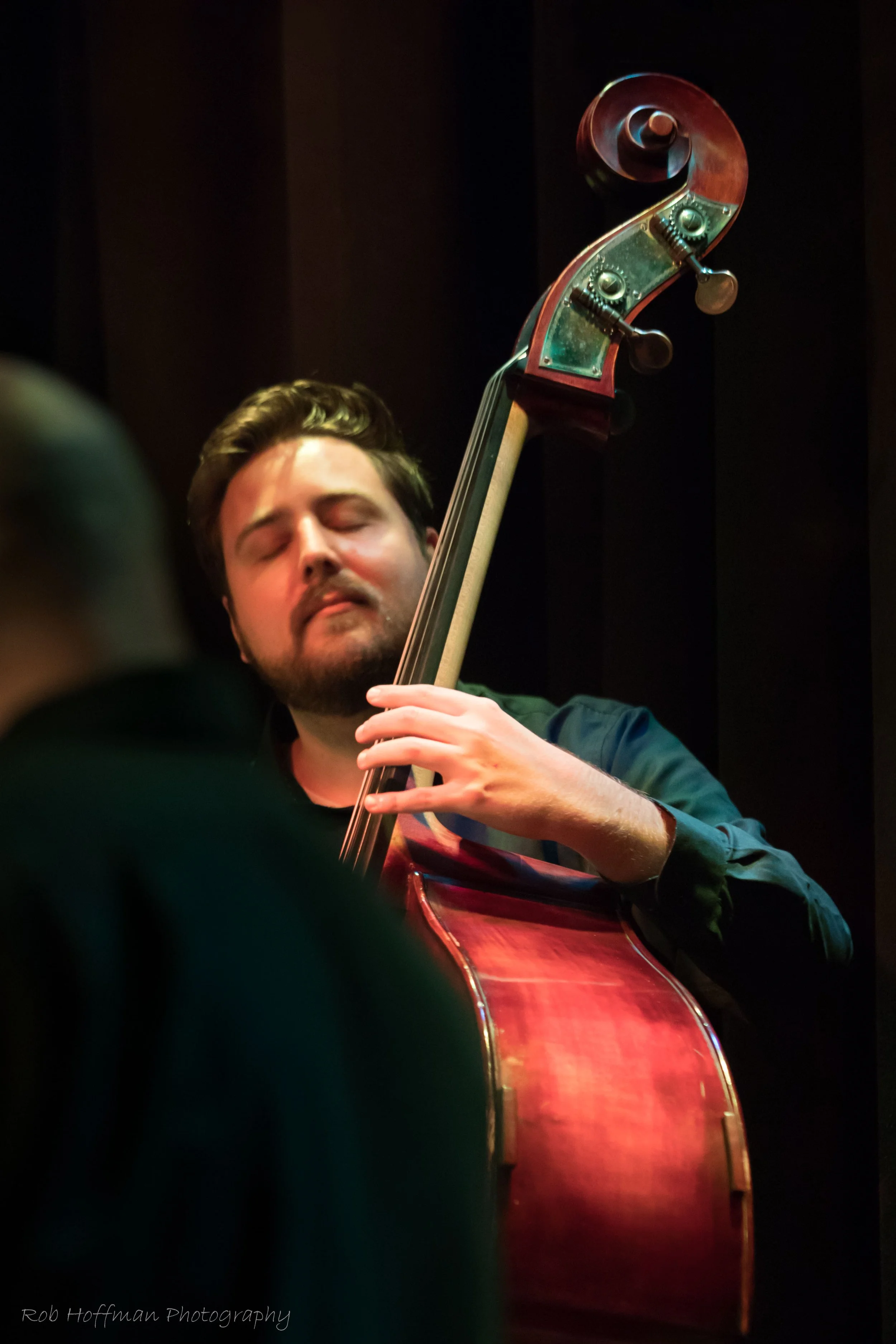 A man passionately playing a double bass with eyes closed, wearing a black shirt, in a dimly lit setting.