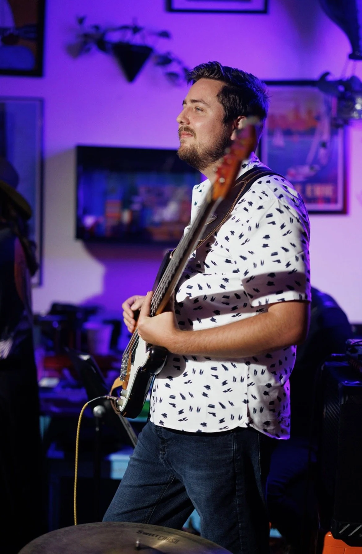 A man playing an electric guitar in a dimly lit room with purple and blue lighting, decorated with framed pictures and wall-mounted plants.