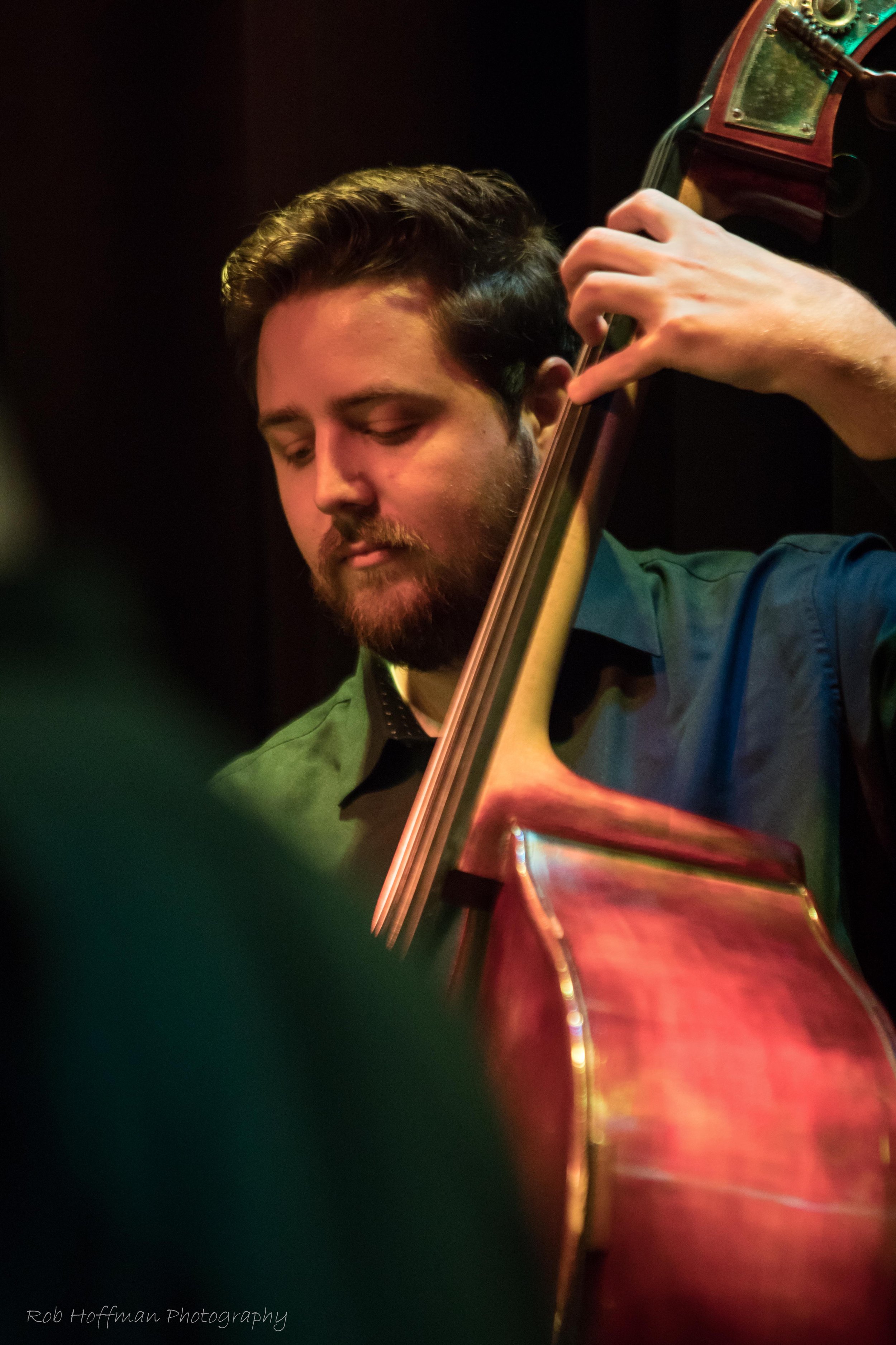 A man with dark hair and a beard playing a double bass, focusing on the instrument with a dark background.