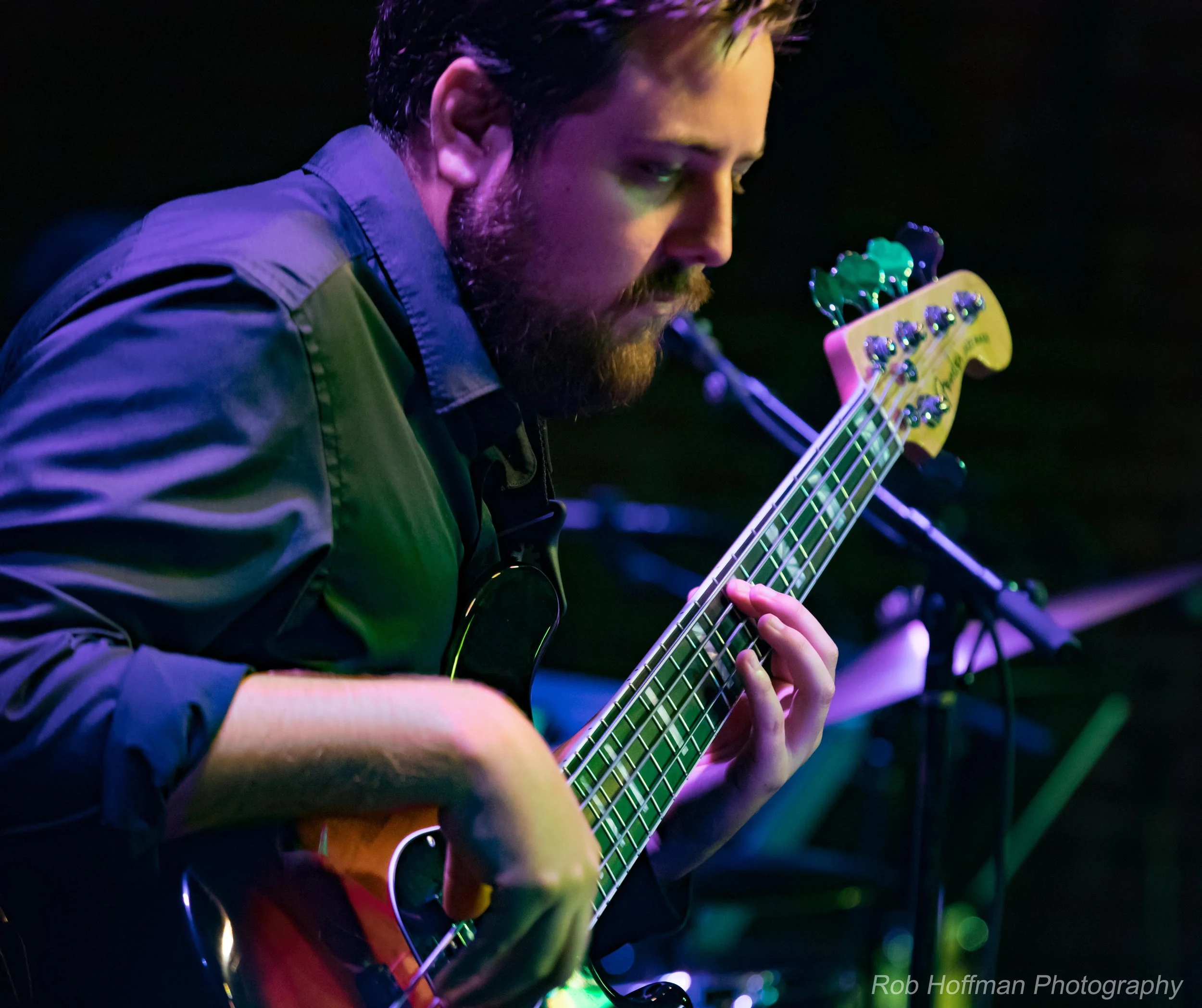 A bearded man playing an electric bass guitar under colorful stage lighting.
