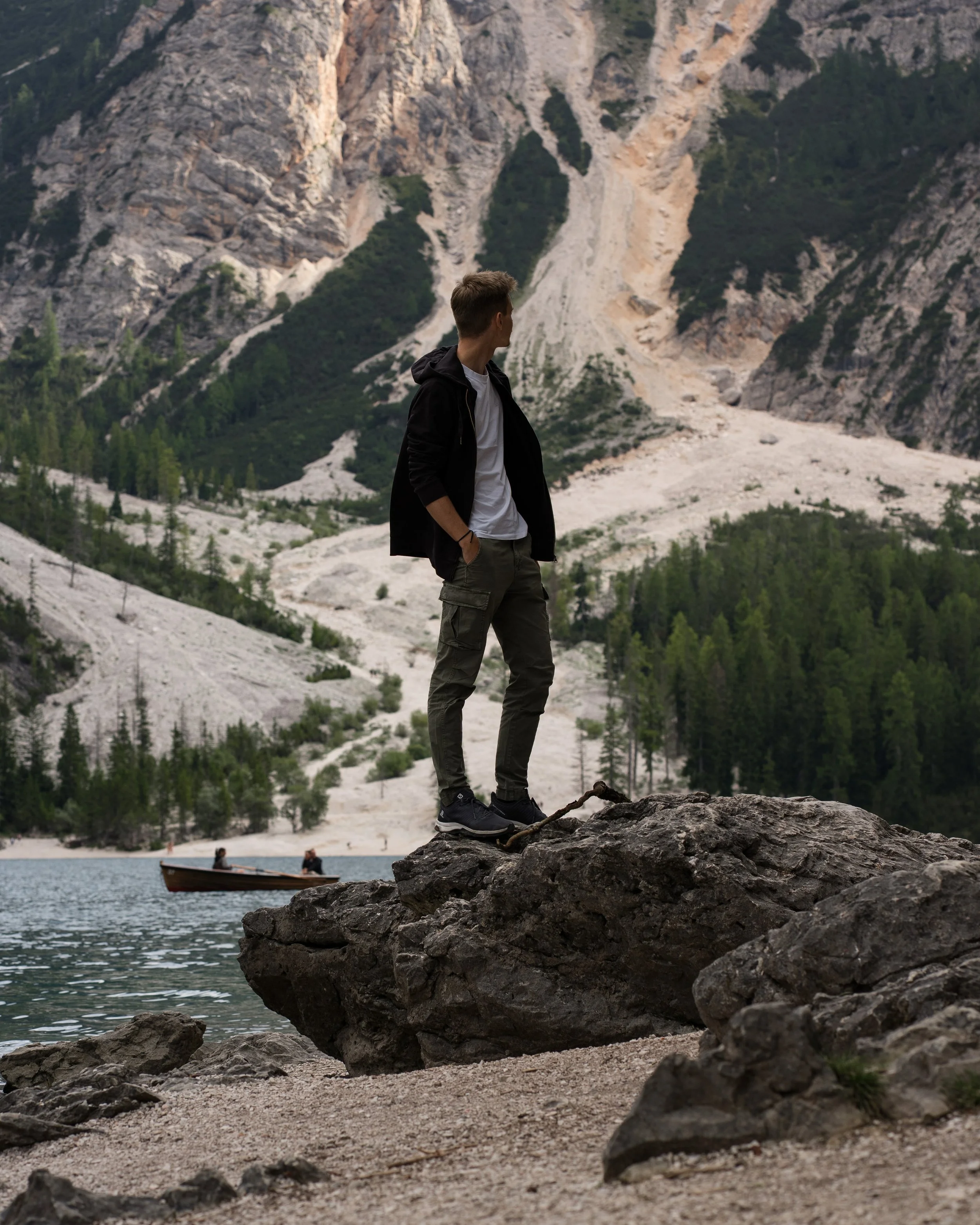A young man standing on a large rock at the edge of a lake with mountains and trees in the background.
