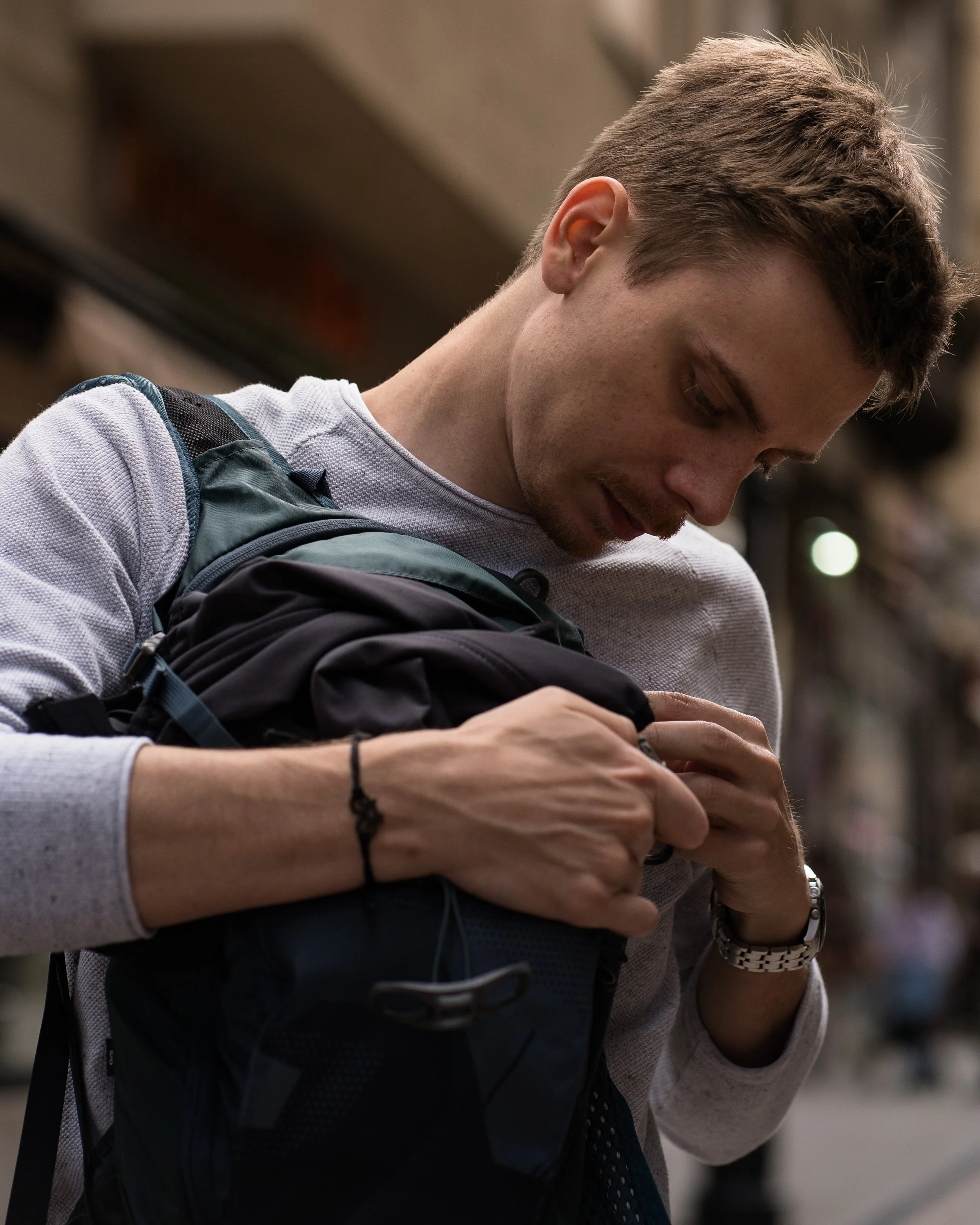 A young man with short hair, wearing a white long sleeve shirt, is looking into and adjusting a backpack while standing outdoors in an urban environment.