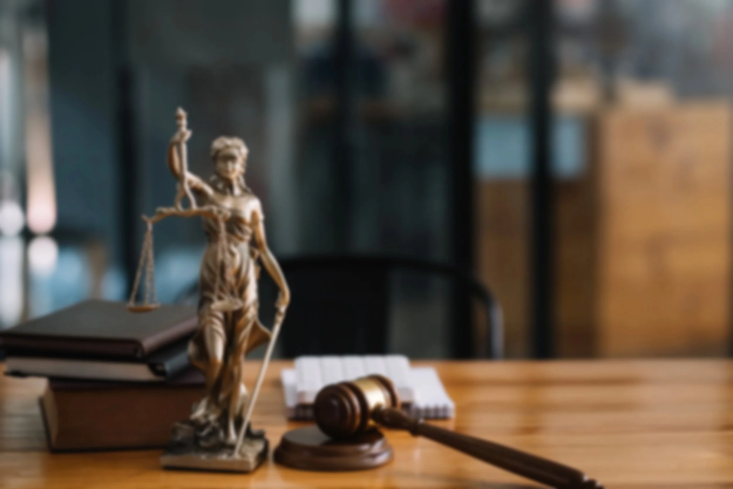 A courtroom scene with a bronze statue of Lady Justice holding scales and a sword on a wooden desk, along with a gavel and legal books, in a blurred background.
