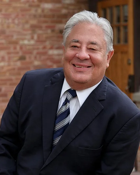 Smiling middle-aged man in a dark suit with a striped tie, standing outdoors in front of a brick building and wooden door.