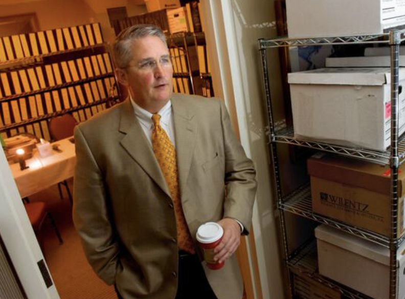 A man in a beige suit and yellow tie holding a coffee cup stands in a room with a table, chairs, and a metal shelving unit filled with boxes.