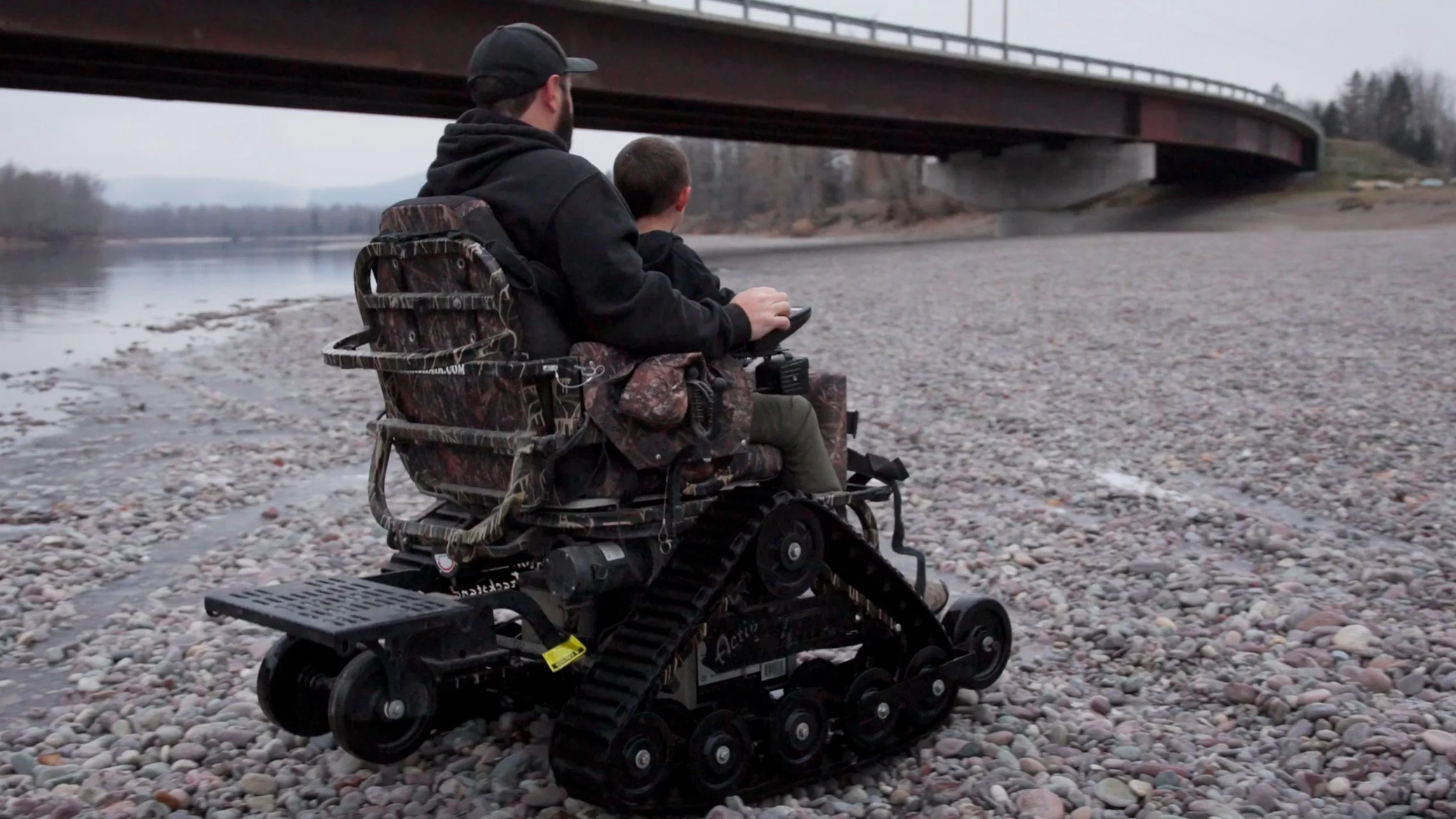 A man and a boy sitting in a tracked wheelchair by a riverbank, under a bridge, with a pebble-covered shoreline.