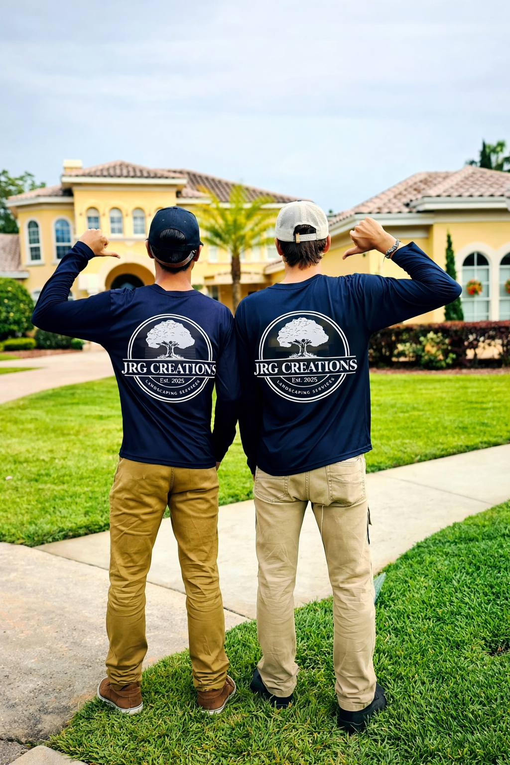 Two men standing on a sidewalk in front of a large house, viewed from the back, wearing matching dark blue shirts with logo, turning and flexing their arms.