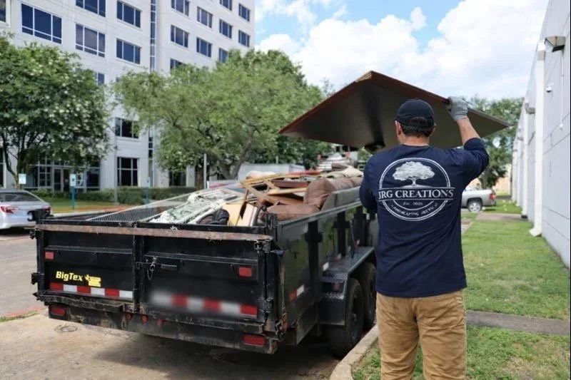 A person wearing a JRG Creations shirt with a logo and tan pants is loading a large piece of furniture onto a black dump trailer filled with furniture and debris, parked outside an apartment complex with trees and other cars.