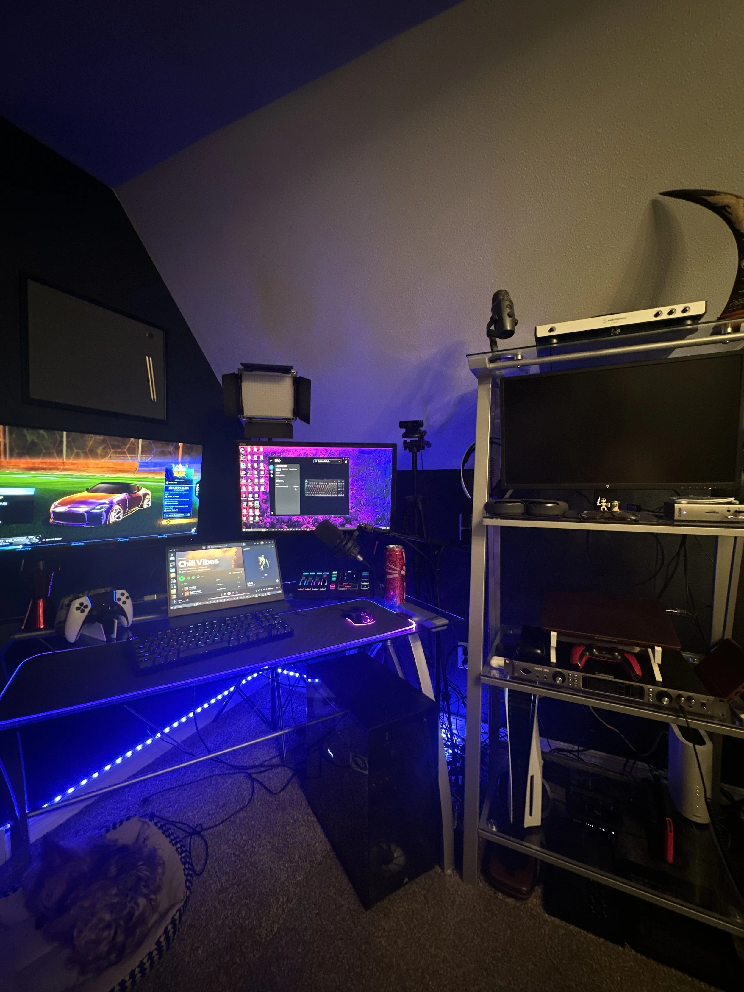A computer gaming setup with multiple monitors, a keyboard, a mouse, a gaming controller, and various electronic devices on a desk and a metal shelving unit, with colorful LED lighting in a dimly lit room.