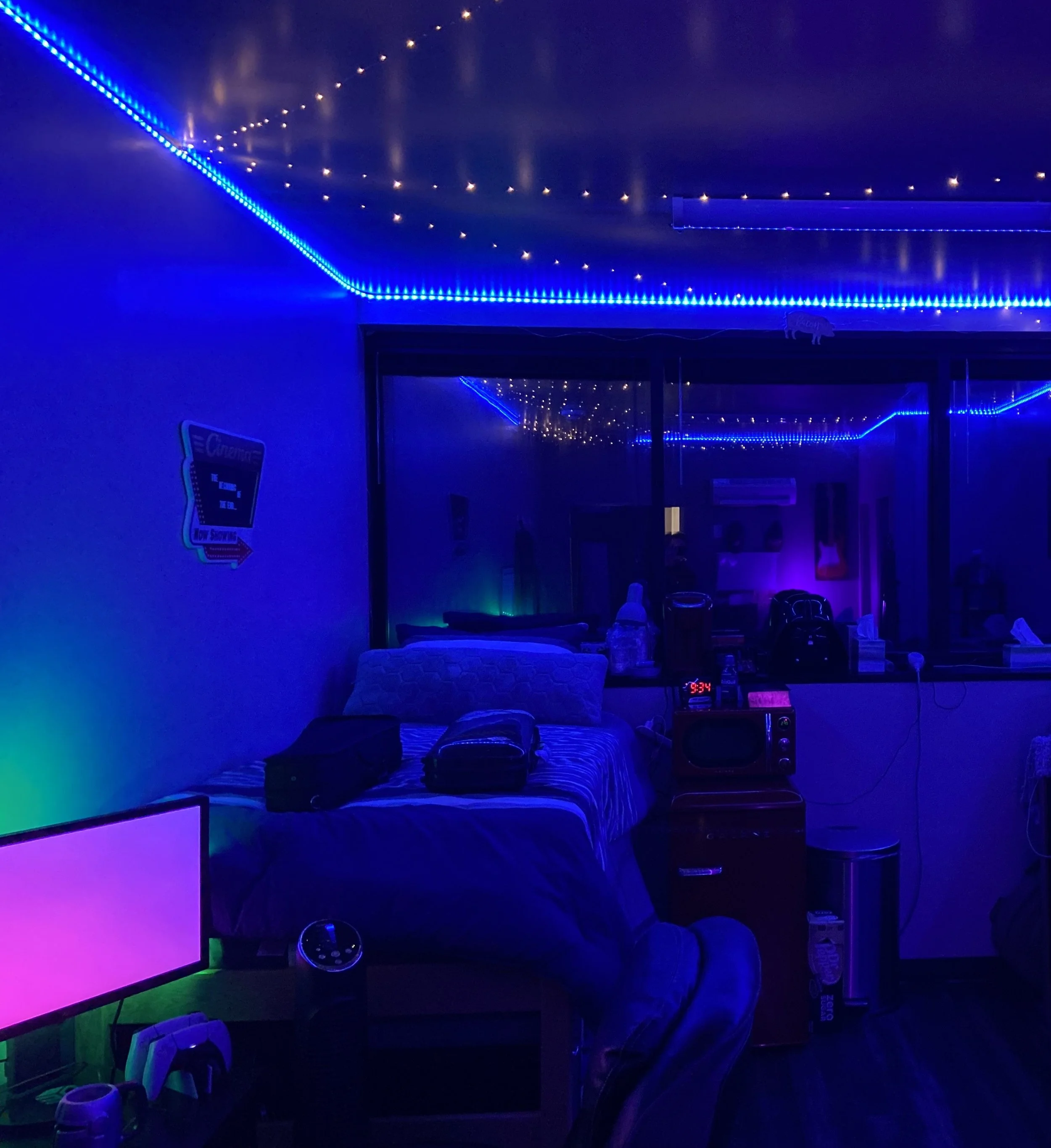 A room decorated with blue string lights along the ceiling, illuminated by some purple and green lighting. There is a bed with pillows and two luggage bags on top, a small dresser with a microwave and a clock, a TV displaying a pink screen, and various electronic devices and decorations.