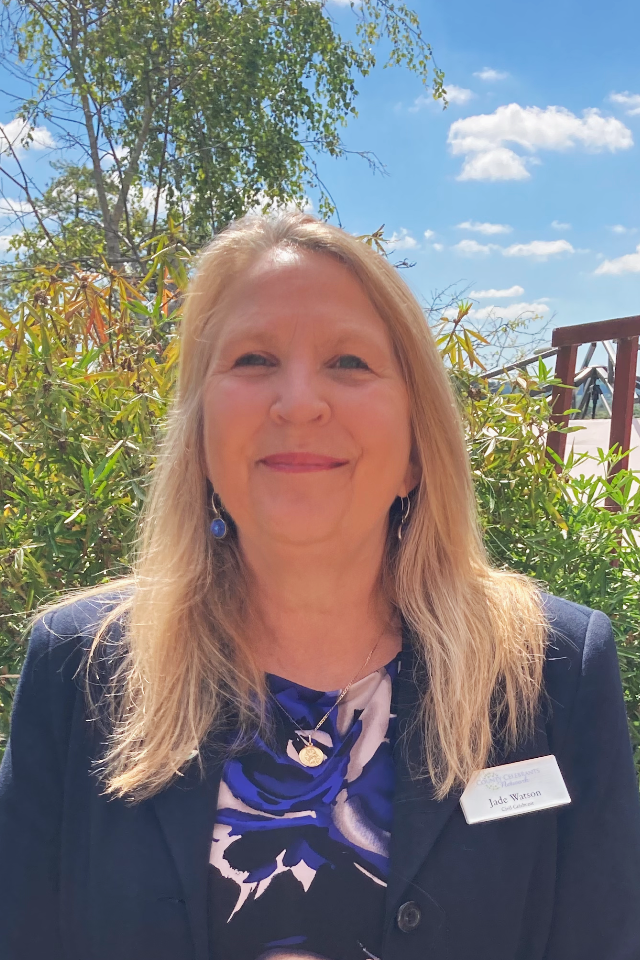 A woman with long blonde hair smiling outdoors on a sunny day, wearing a navy blazer and a patterned blouse, with a name tag reading 'Jade Watson'. She is standing in front of green shrubs and a blue sky with clouds.