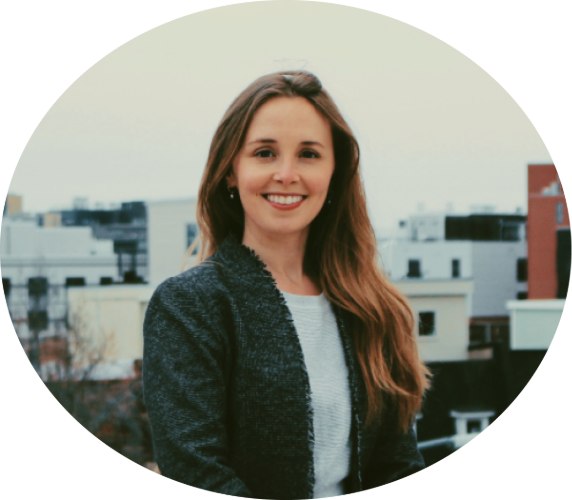 A woman with long brown hair smiling outdoors in an urban setting with buildings in the background.