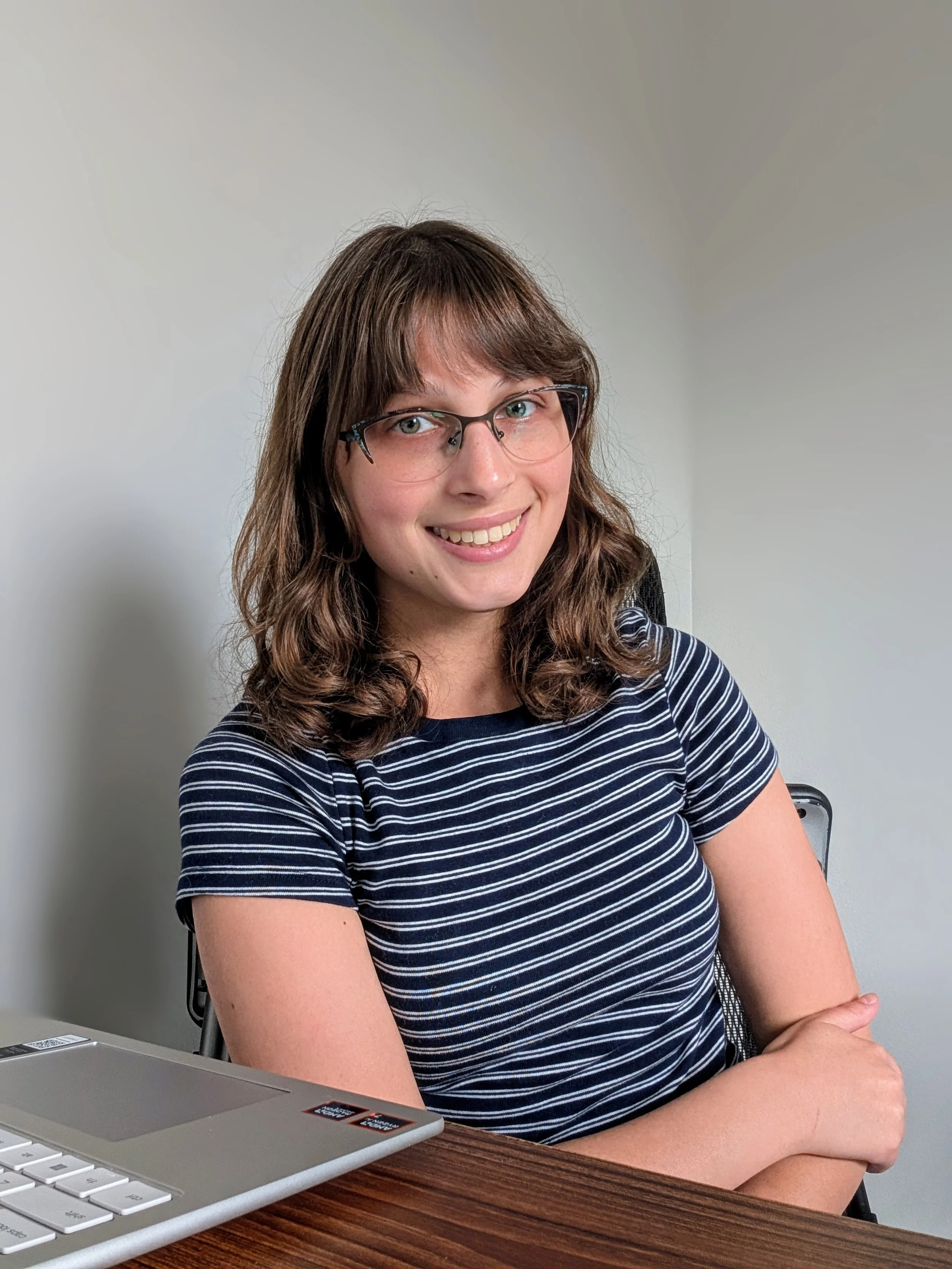 A woman with brown hair, glasses, and a striped shirt smiling and sitting at a desk, arm crossed, with a laptop nearby.