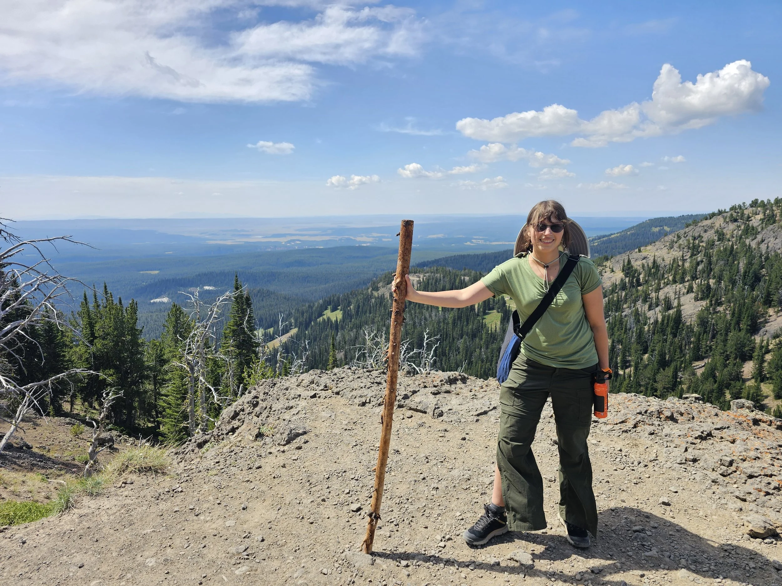 A woman with sunglasses smiling and holding a walking stick on a mountain trail, with scenic views of forests, mountains, and a blue sky with scattered clouds in the background.