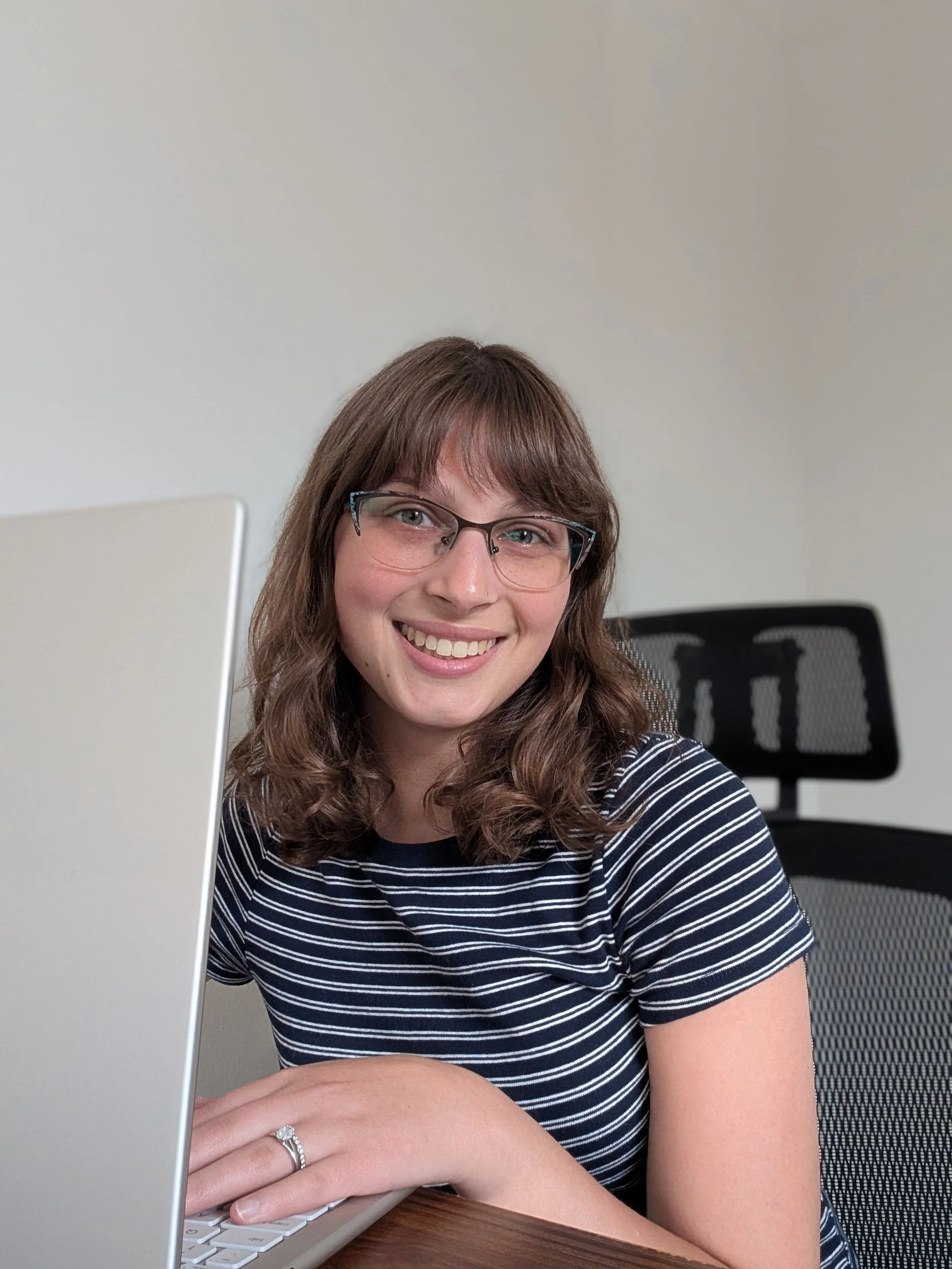 A woman with brown hair, glasses, and a striped shirt smiling while working on a laptop in an office setting.