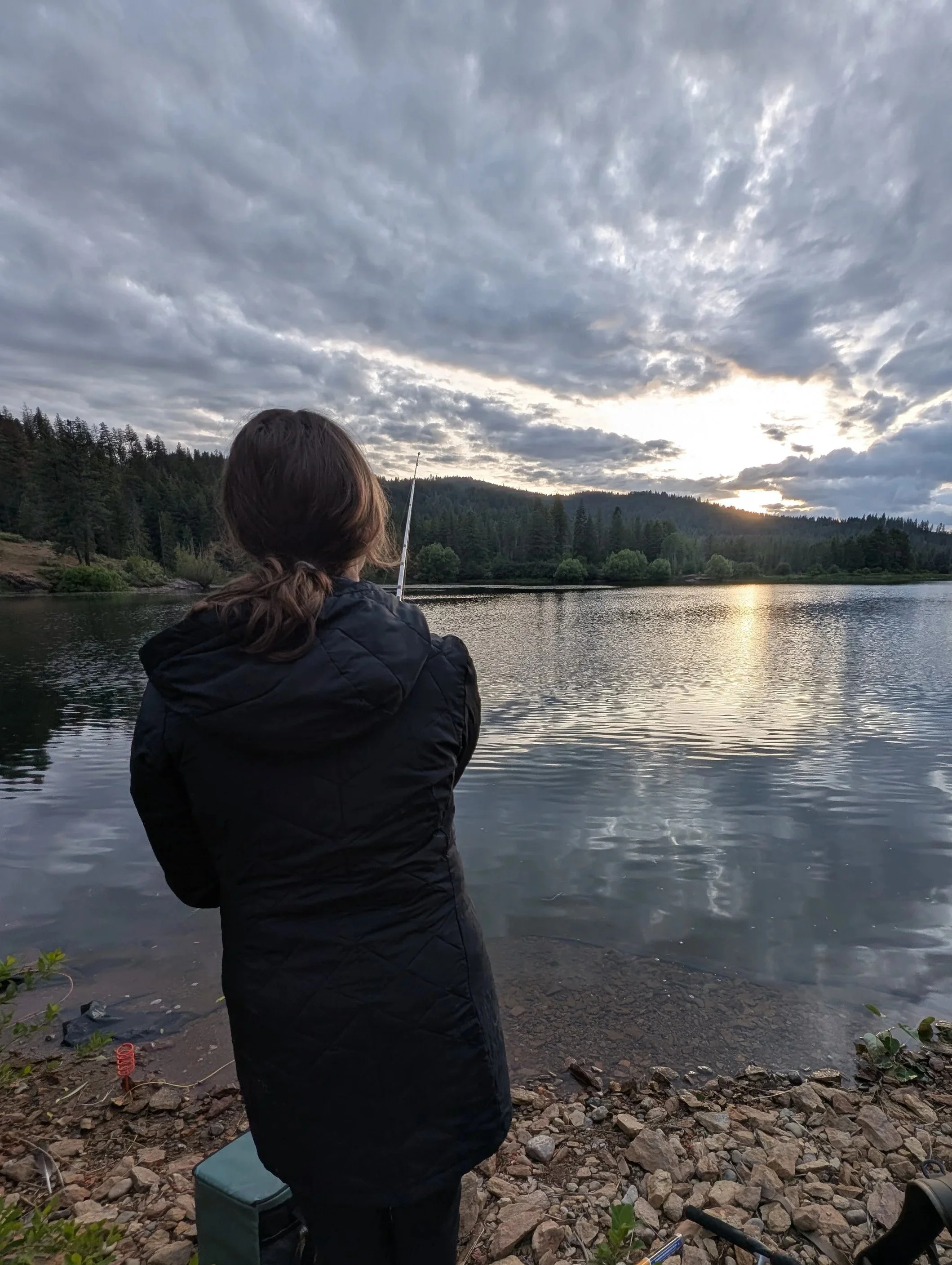 Person in a black jacket fishing by a river with a cloudy sky and forested hills in the background during sunset.