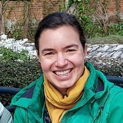 A woman smiling outdoors with greenery and a rocky background.