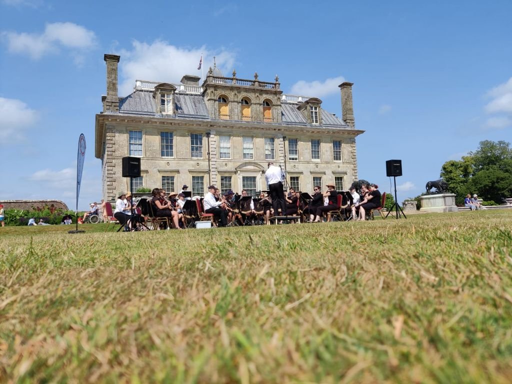 An outdoor orchestra performing in front of a historic mansion with a blue sky and some clouds.