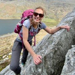 Woman with sunglasses climbing a gray rock outdoors, carrying a pink backpack and smiling.