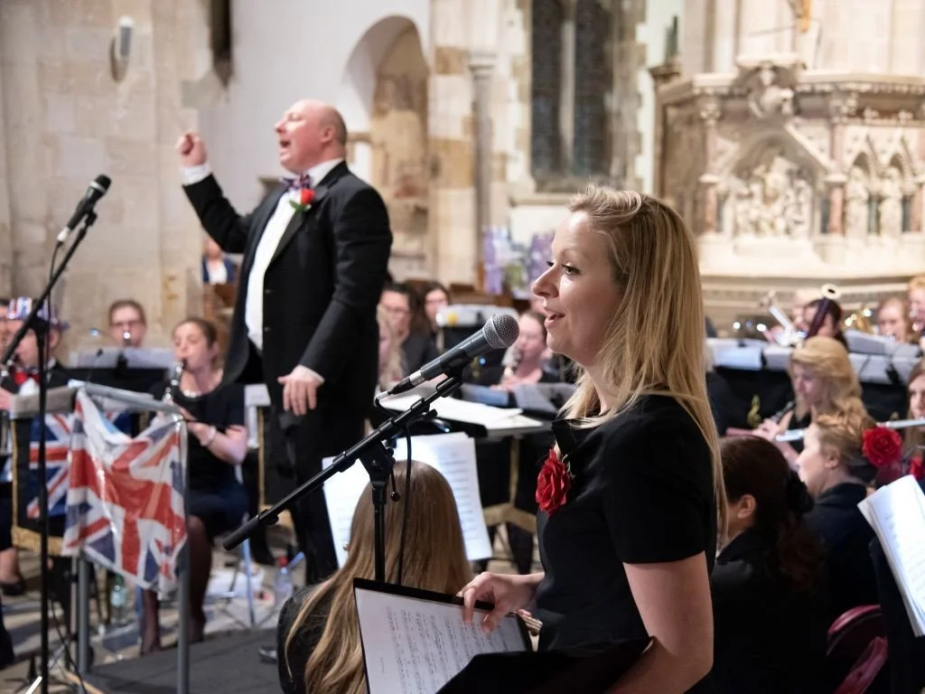 Woman singing into microphone during a concert next to conductor and with band behind