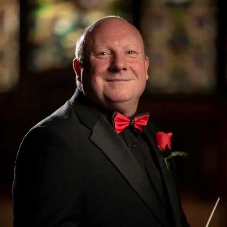 A man in a tuxedo with a red bow tie and a red pocket square, smiling in a dimly lit setting.