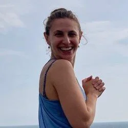A woman smiling outdoors against a cloudy sky, wearing a blue tank top.