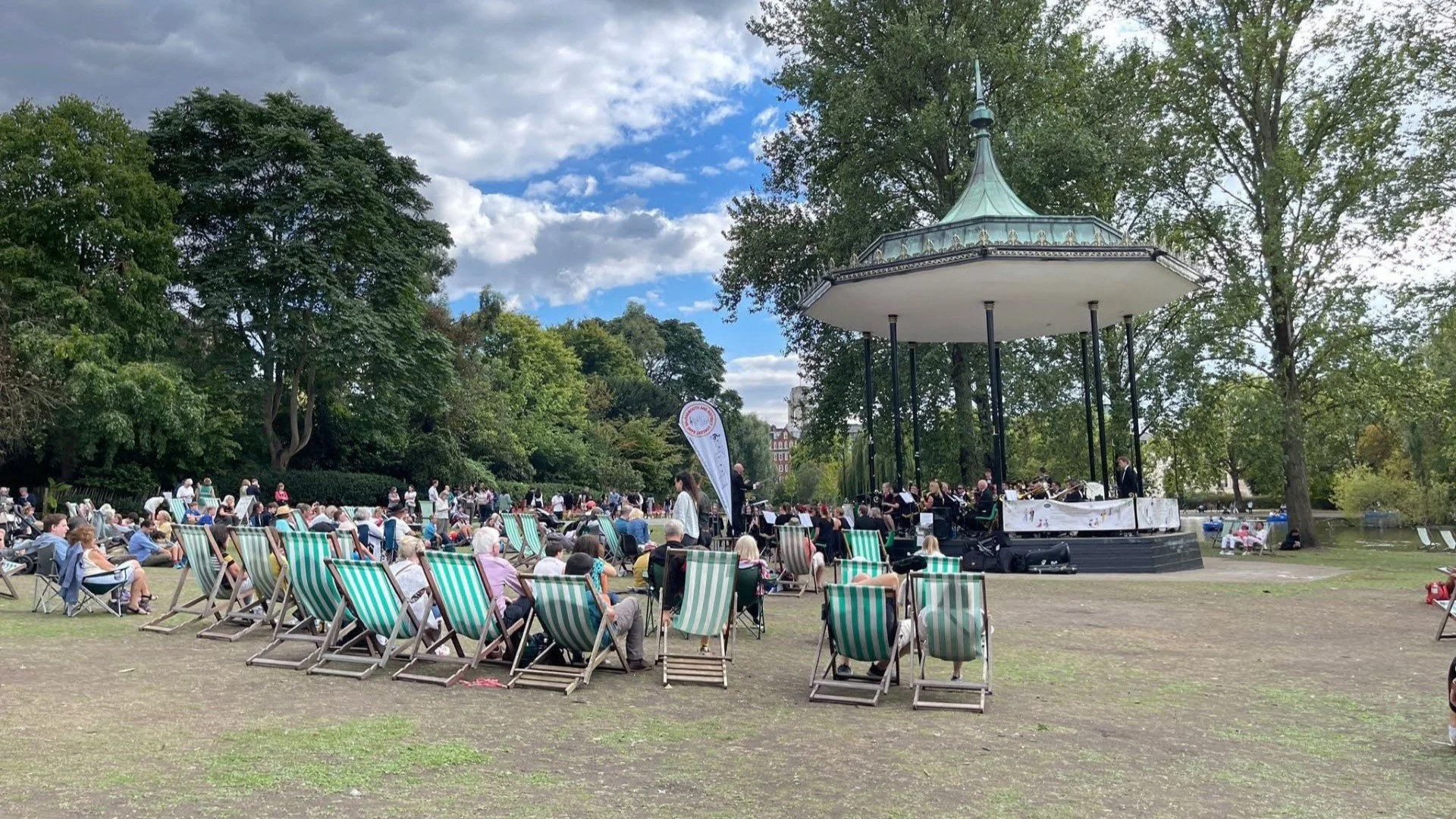 Band performing at Regent's Park Bandstand in London with audience sitting in deck chairs watching