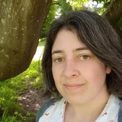 A woman with dark hair and light skin outdoors next to a tree, smiling gently at the camera.