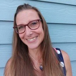 A woman with long hair wearing glasses and a sleeveless dress, smiling in front of a blue wooden wall.