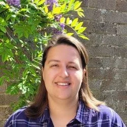 A woman with shoulder-length brown hair smiling outdoors, standing in front of green foliage and a brick wall
