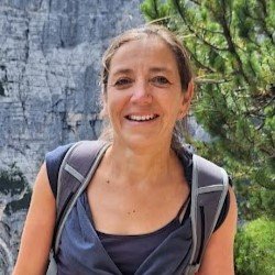 Smiling woman with brown hair tied back, wearing a gray tank top and backpack, outdoors near rocky cliffs and green trees.