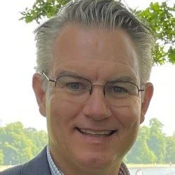 A middle-aged man with gray hair wearing glasses, smiling outdoors with trees and a lake in the background.