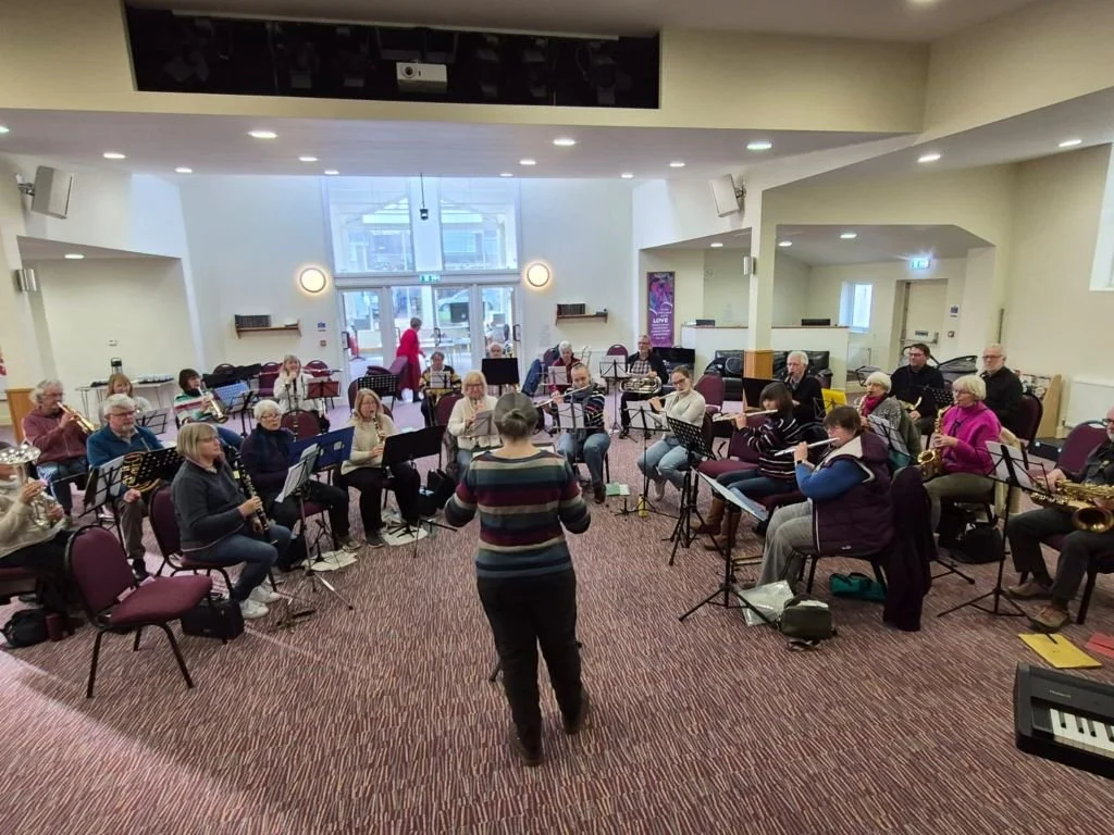 A large group of musicians playing wind instruments in a concert hall, directed by a conductor, with some people seated in the background near the entrance.