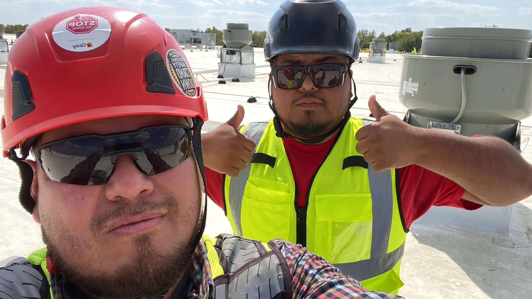 Two construction workers on a rooftop wearing safety helmets, sunglasses, and high visibility vests; one taking a selfie while the other gives a thumbs-up.