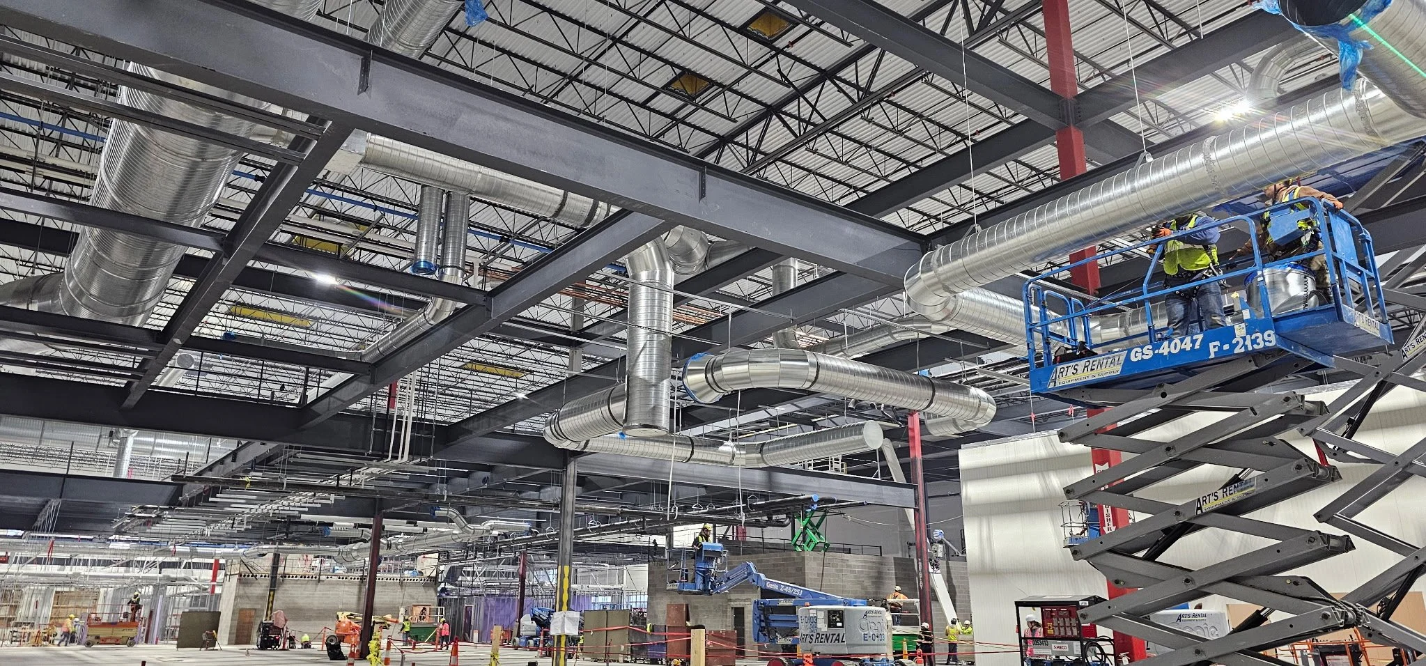 Construction workers installing large metallic HVAC ducts in a building under construction, with scaffolding and equipment visible.