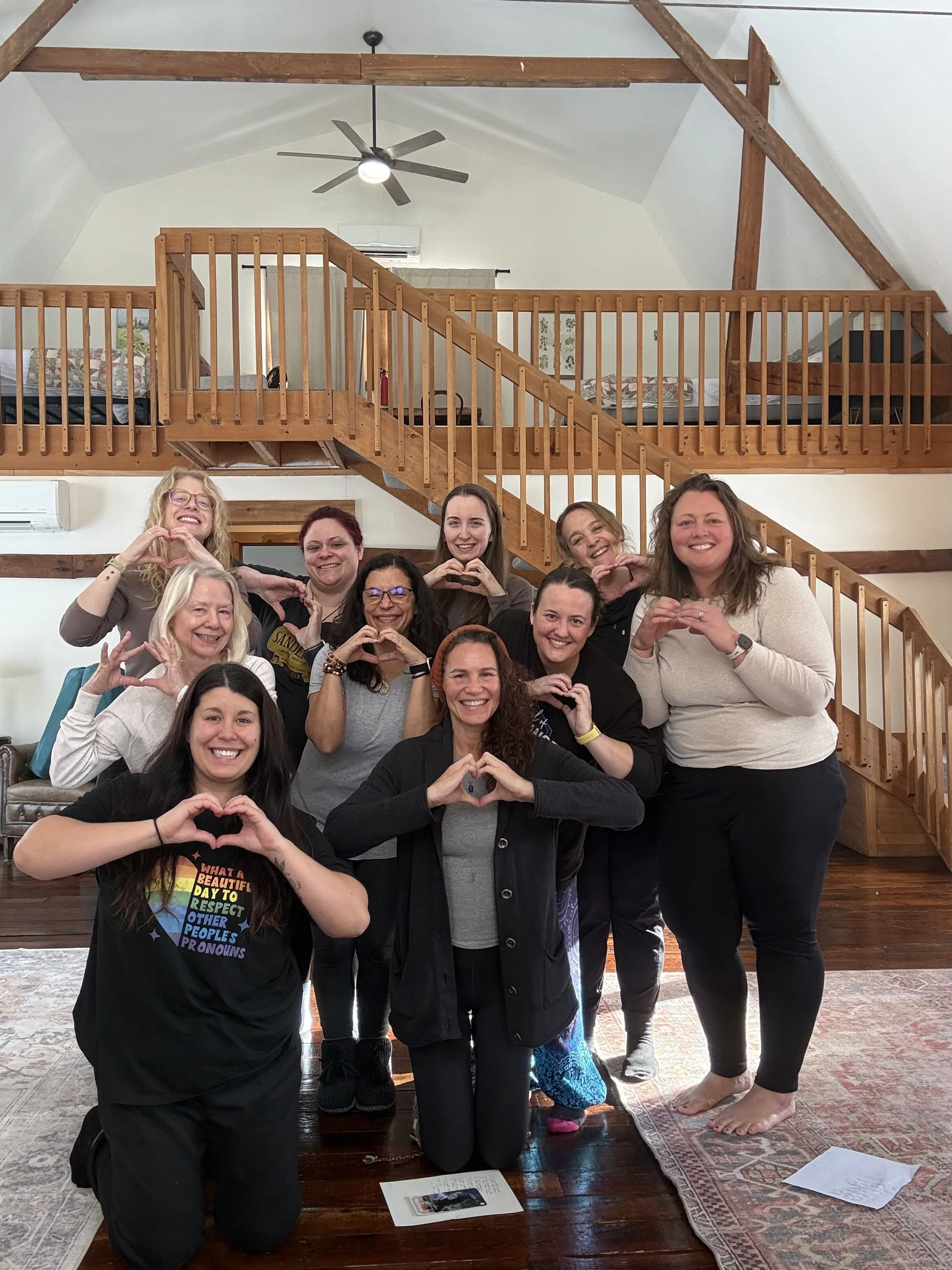 Group of ten women smiling and making heart shapes with their hands inside a wooden-floored room with a high ceiling, wooden beams, and a balcony in the background.