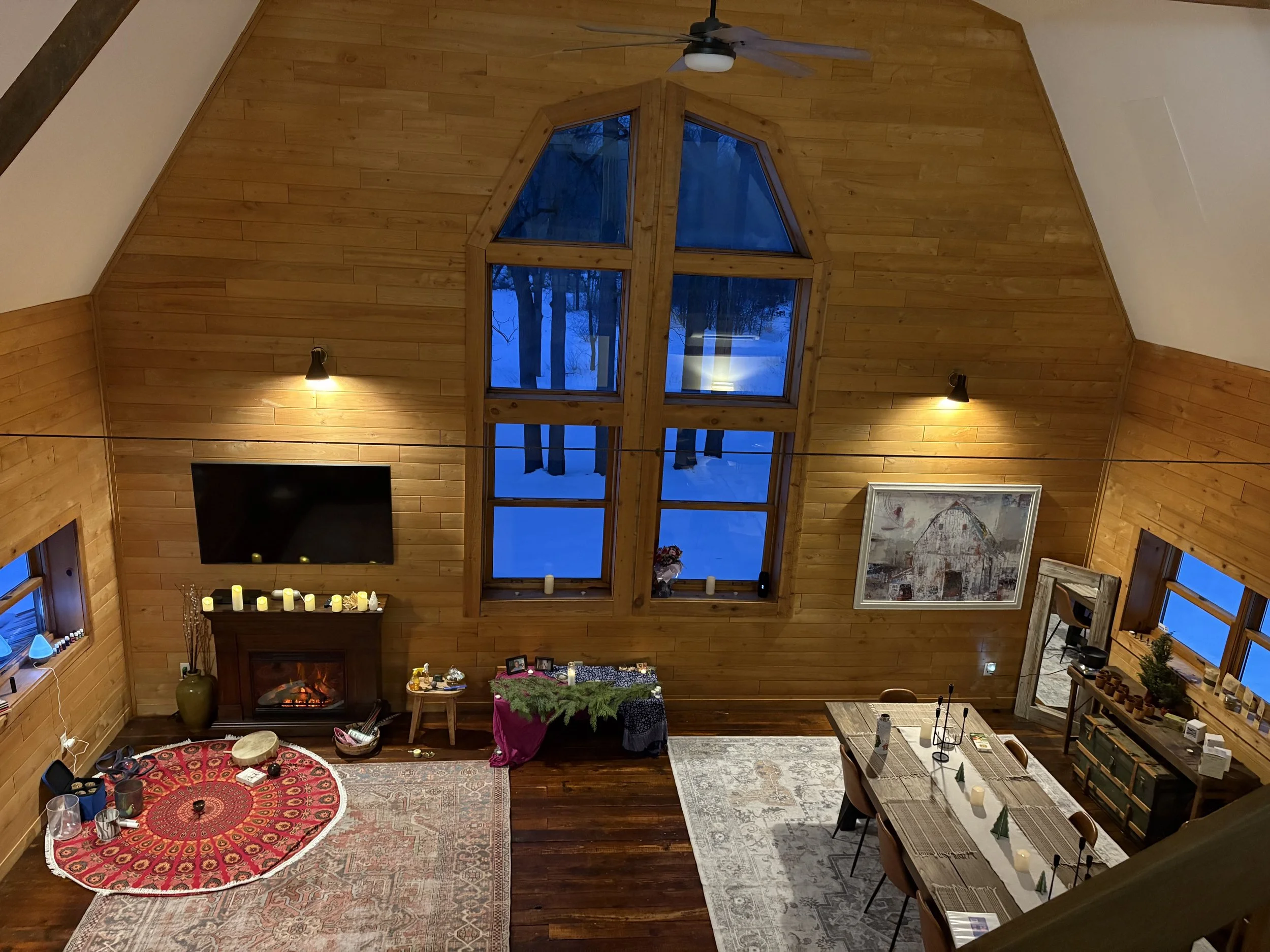 Living room in a wooden cabin with high ceiling, large front window, fireplace, TV, dining table, and holiday decorations.