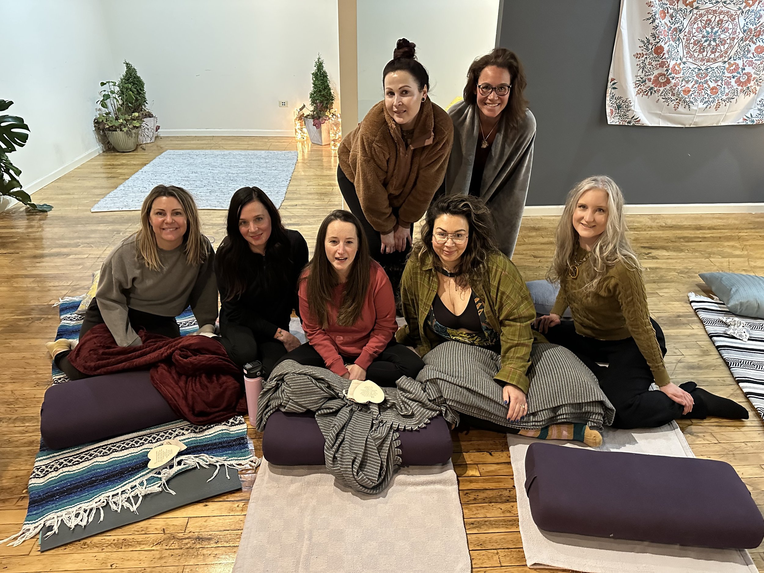 Seven women sitting and standing on the wooden floor of a cozy room with meditation cushions, blankets, and a tapestry on the wall, participating in a group activity or gathering.