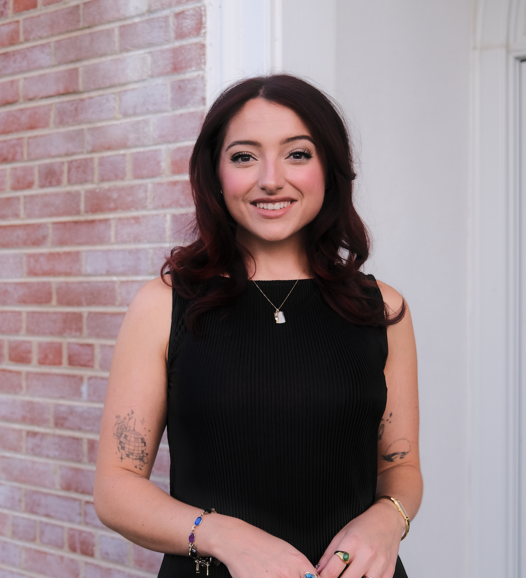 A young woman with dark red hair, wearing a black sleeveless top, standing outside near a brick wall and a white building, smiling at the camera.