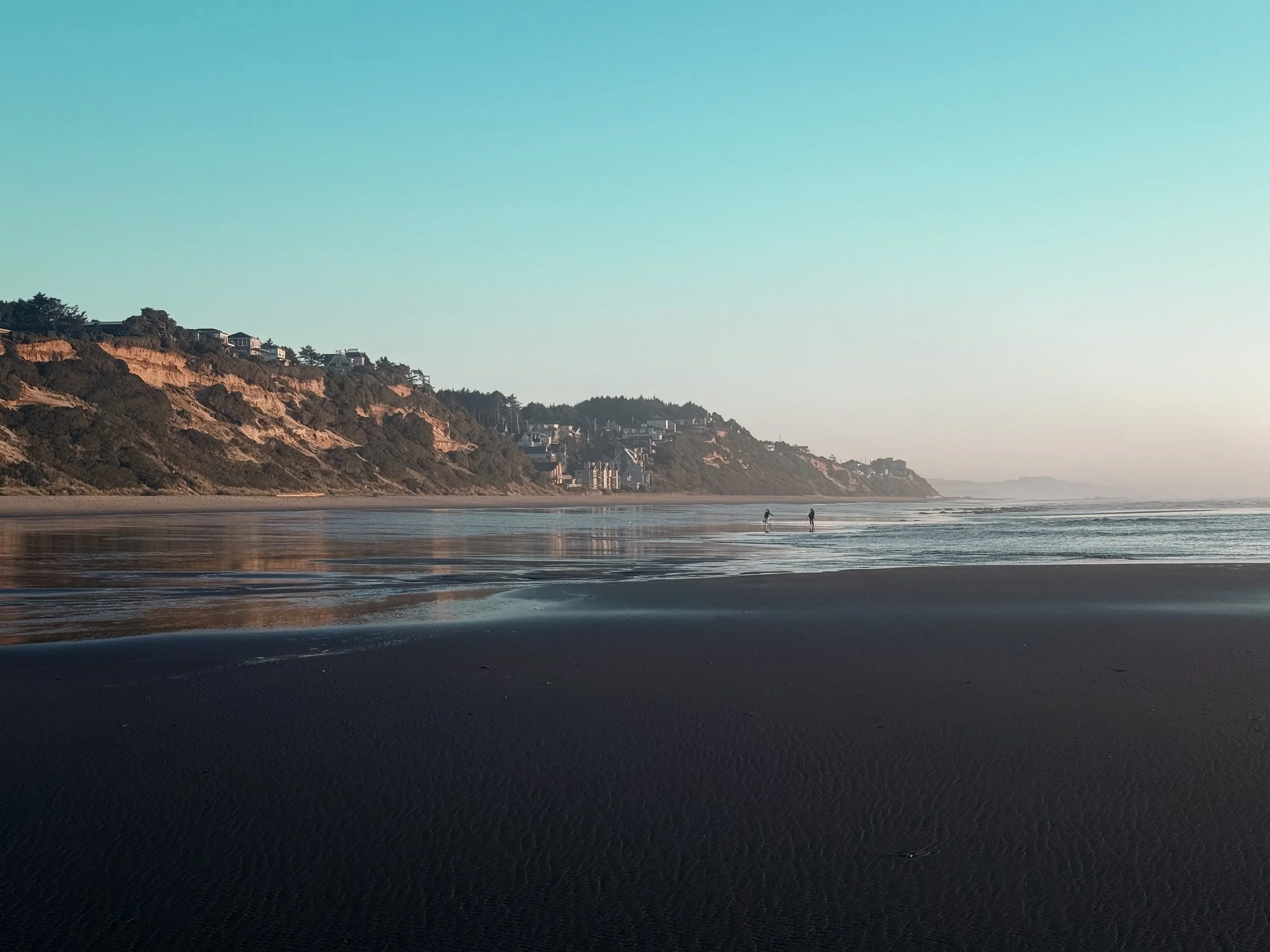 A peaceful beach scene with two people walking along the shoreline, sandy beach, waves, cliffs with houses in the background, and a clear sky.